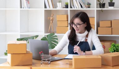 Young beautiful happy asian business woman owner of SME online using laptop receive order from customer with parcel box packaging at her startup home office, online business seller and deliver