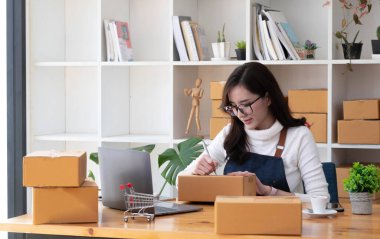 Young beautiful happy asian business woman owner of SME online using laptop receive order from customer with parcel box packaging at her startup home office, online business seller and deliver
