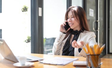 Ofiste akıllı telefon ve dizüstü bilgisayar kullanan mutlu Asyalı genç iş kadını.