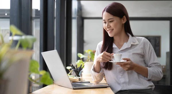 Beautiful young smiling Asian businesswoman working on laptop and drinking coffee, Asia businesswoman working document finance and calculator in her office