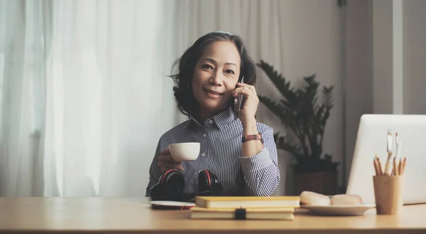 Happy pretty mature woman speaking on mobile phone portrait. Senior grey haired lady making telephone call, sitting on couch at home, enjoying talk, communication, looking at camera, smilin