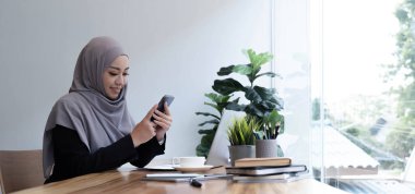 Mor başörtüsü takmış, yeni cep telefonu kullanan, fotokopi alanına bakan ve gülümseyen, iş için en yeni mobil uygulamayı kullanan güzel, genç ve güzel bir kadın.
