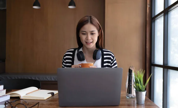 Calm and relaxed young Asian woman sipping a morning coffee while reading an online content on her portable laptop at the desk