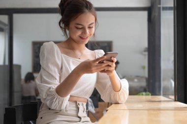 Kafede akıllı telefon kullanan Asyalı genç bir kadın. Evde akıllı telefon kullanan, mesajlaşan ya da sosyal ağları karıştıran gülümseyen bir kadın