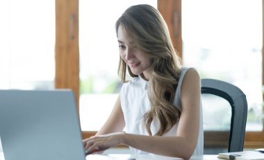 Ofisinde iş yerinde oturan mutlu Asyalı genç iş kadını. Ofiste dizüstü bilgisayarda çalışan genç bir kadın.