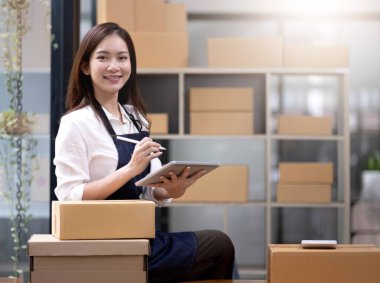 Genç, çekici Asyalı kadın şirket sahibi kameraya bakın. Evde kutuyla mutlu bir şekilde çalışıyor. Paket teslimatını sme tedarik zincirinde hazırlayın..