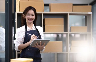 Genç, çekici Asyalı kadın şirket sahibi kameraya bakın. Evde kutuyla mutlu bir şekilde çalışıyor. Paket teslimatını sme tedarik zincirinde hazırlayın..