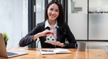 Miniature house in the hands of an Asian woman real estate agent home loan working at the office. Looking at the camera