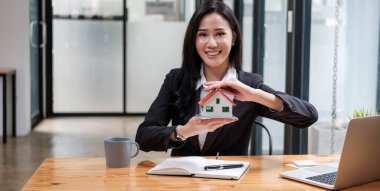 Miniature house in the hands of an Asian woman real estate agent home loan working at the office. Looking at the camera