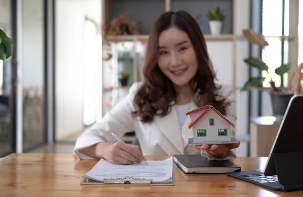 Miniature house in the hands of an Asian woman real estate agent home loan working at the office. Looking at the camera