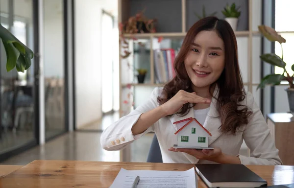 Miniature house in the hands of an Asian woman real estate agent home loan working at the office. Looking at the camera