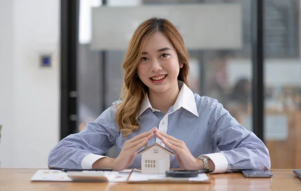 Miniature house in the hands of an Asian woman real estate agent home loan working at the office. Looking at the camera