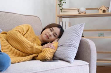 beautiful asian woman in sweater sleeping on a couch at home with mobile phone in home