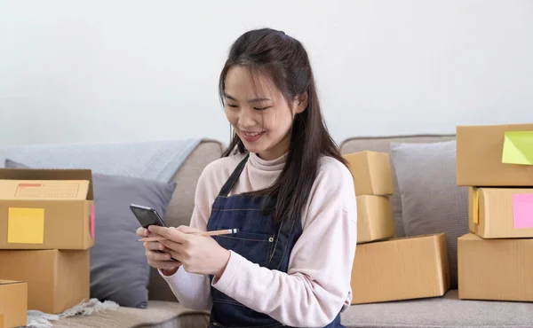 Happy young Asian woman entrepreneur, Smile for sales success after checking order from online shopping store in a smartphone at home office, Concept of merchant business online and eCommerc