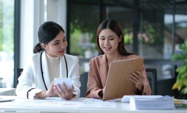 İki genç Asyalı iş kadını yatırım projesinin çalışma ve planlama stratejisini tartıştı. İş adamları ofiste dizüstü bilgisayarla konuşuyor.