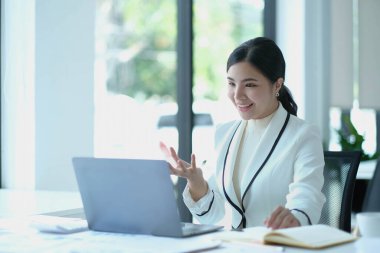 Distant communication. Friendly pretty asian brunette girl, manager, freelancer, sit in the kitchen, talking on video call by laptop with the client or employees, discussing project, smilin