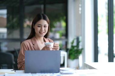 Smiling young Asian businesswoman holding a coffee mug and laptop at the office.