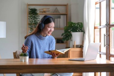 Gülümseyen Asyalı genç kız kulaklık takıyor ve laptoptan arıyor. Mutlu güzel kız öğrenci bilgisayar ekranına bakıyor webinar izliyor ya da webcam 'den görüntülü sohbet yapıyor.