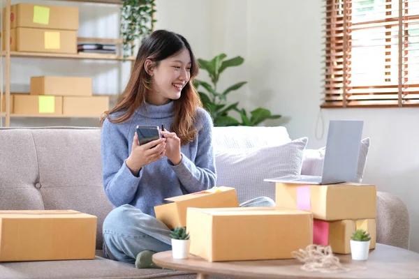 Young beautiful happy asian business woman owner of SME online using laptop and smartphone receive order from customer with parcel box packaging at her startup home office, online business seller and deliver