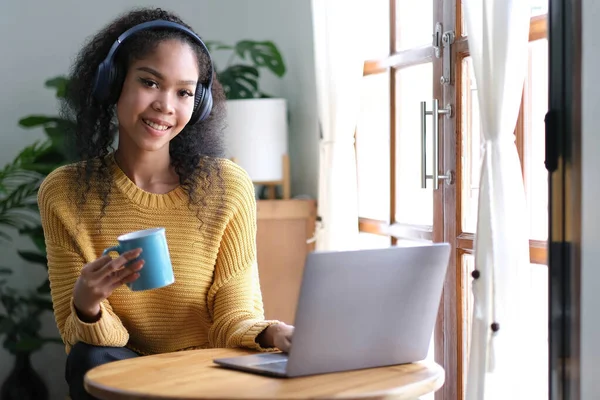 Smiling young asian woman using laptop web camera while holding a cup of coffee and wearing headphones in theliving room at home.