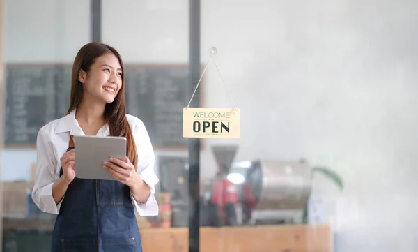 Asian Happy business woman is a waitress in an apron, the owner of the cafe stands at the door with a sign Open waiting for customers. Small business concept, cafes, and restaurant