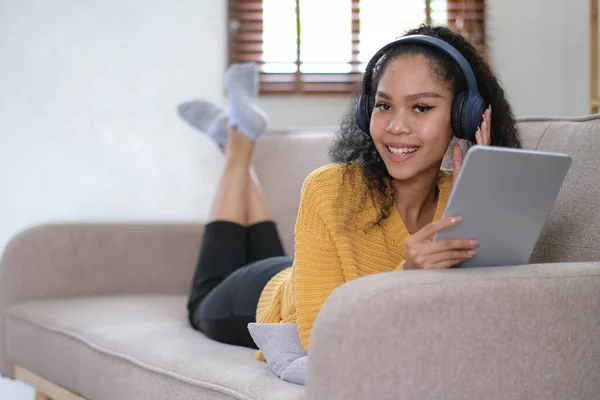 african young woman relaxing at home lying on sofa and listening to music on tablet wearing headphones. girl relaxing on the sofa of a cozy hom