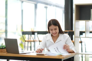 Gülümseyen Asyalı iş kadını, tabloyu ve grafiği analiz ederek piyasadaki değişiklikleri gösteriyor ve ofiste hesap makinesi kullanıyor.