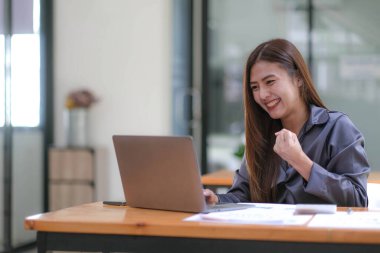 Mutlu Asyalı başarılı iş kadını iş yerinde dizüstü bilgisayarlı akıllı telefonla zafer kazanıyor.