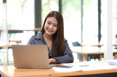 Güzel Asyalı iş kadını laptopunu çalışmak ve çalışmaktan zevk almak için kullanıyor, not alıyor, ödevi gözden geçiriyor ve ofiste gülümsüyor.