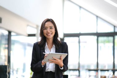 Çekici Asyalı kadın gülümseyerek elinde tabletle ofisteki kameraya bakıyor.