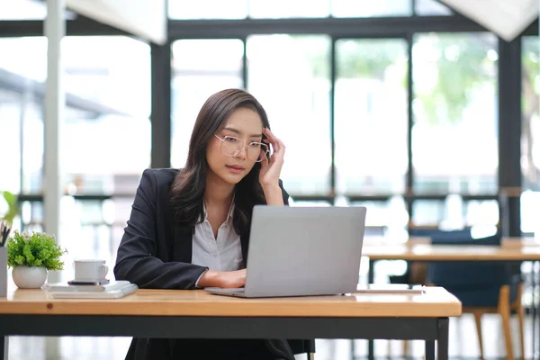 Exhausted businesswoman working at office desk with a laptop and touching her forehead, she is having an headache, stressful job concept.