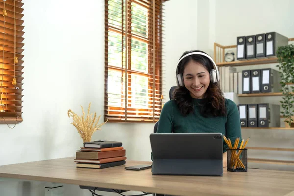 Smiling woman wearing wireless headphones working typing on notebook sit at desk in office workplace. Enjoy e-learning process, easy comfortable application usage, listen music during workday concept.