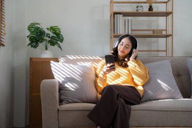 Gülümseyen genç bir kadın evdeki kanepede oturup kulaklıkla akıllı telefondan video konuşması yapıyor. Kulaklıklı mutlu bin yıllık kadın cep telefonu kullanıyor. Web kamerası dijital iletişimi var...