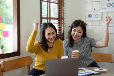 İki genç Asyalı iş kadını, modern bir ofiste dizüstü bilgisayarla mutlu bir şekilde gülümseyerek iş yerinde neşeli bir ifade sergiliyorlar...