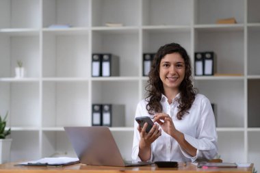 Genç gülümseyen iş kadın ofiste, bilgisayarın yakınında Smartphone kullanarak kopyalama alanı..