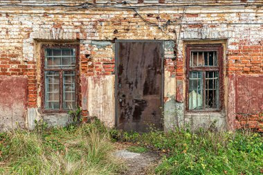 Yıpranmış tuğlalı eski bir duvar, iki parmaklıklı pencere ve kilitli paslı bir demir kapı.