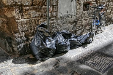 Siyah plastik çöp torbaları ve arka sokakta eski dokulu taş bir duvar ve rögar kapağı olan eski bir bisiklet.