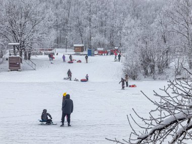Pazar günü, ebeveynler ve çocuklar şehir parkındaki donmuş bir göletin eğimli sahilinden borulara biniyorlar.