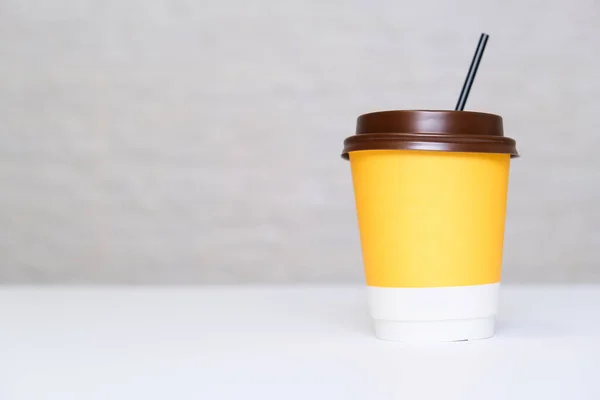 A yellow paper disposable takeout coffee cup with a brown cap and a straw on white background against light brick wall with copy space.
