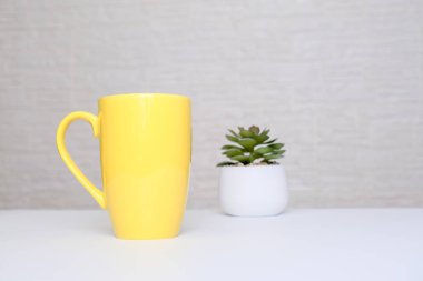 Büyük sarı seramik kupa, beyaz bir masanın üzerinde bir fincan Amerikan kahvesi. Fotokopi alanı olan tuğla duvara karşı saksı bitkisi, ofis kahvesi konsepti.
