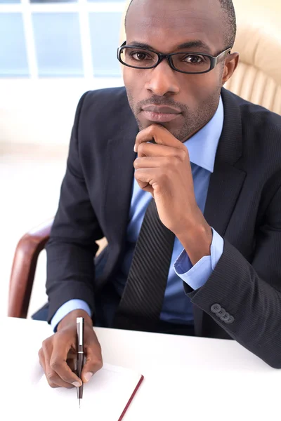 Empresario africano escribiendo en su bloc de notas y tomándose de la mano en la barbilla —  Fotos de Stock