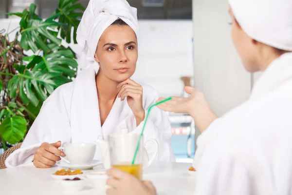 Spending time at spa. Two beautiful young women in bathrobe drinking tea and talking to each other while sitting in front of swimming pool — Stock Photo, Image