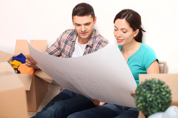 Examinando un plano. Hermosa pareja joven sentada en el suelo y mirando el plano mientras cajas de cartón que yacen alrededor de ellos — Foto de Stock