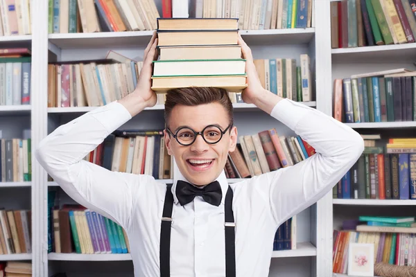 Jovem alegre na biblioteca e segurando uma pilha de livros na cabeça — Fotografia de Stock