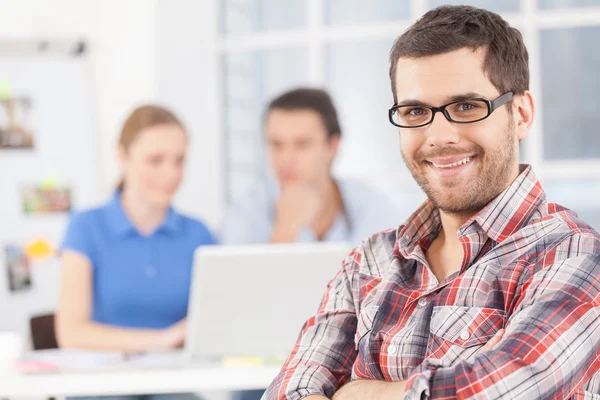 Confiando en su equipo. Joven alegre en gafas mirando a la cámara y sonriendo mientras sus colegas trabajan en el fondo —  Fotos de Stock