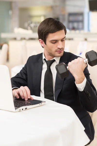 Hombre en ropa formal sentado en el ordenador portátil y sosteniendo una mancuerna —  Fotos de Stock