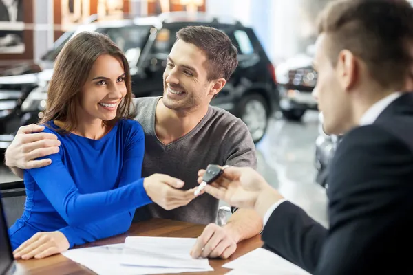 Vendedor de coches dando la llave del coche nuevo a los propietarios atractivos jóvenes — Foto de Stock