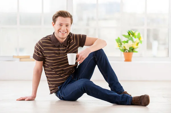 Homem segurando uma xícara e sorrindo para a câmera enquanto sentado no chão — Fotografia de Stock