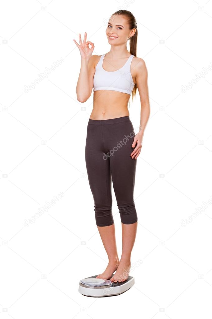 Woman in sports clothing standing on weight scale and showing OK sign