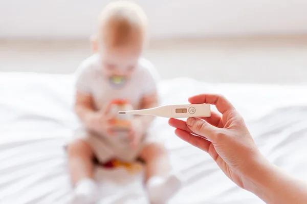 Hand holding thermometer while baby sitting on the bed — Stock Photo, Image